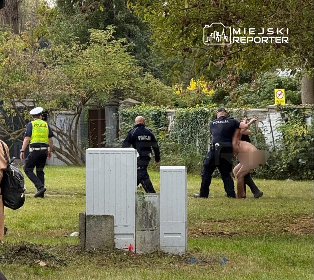 Policjanci zatrzymują nagiego mężczyznę na trawniku przy ogrodzeniu i zarośniętym terenie.