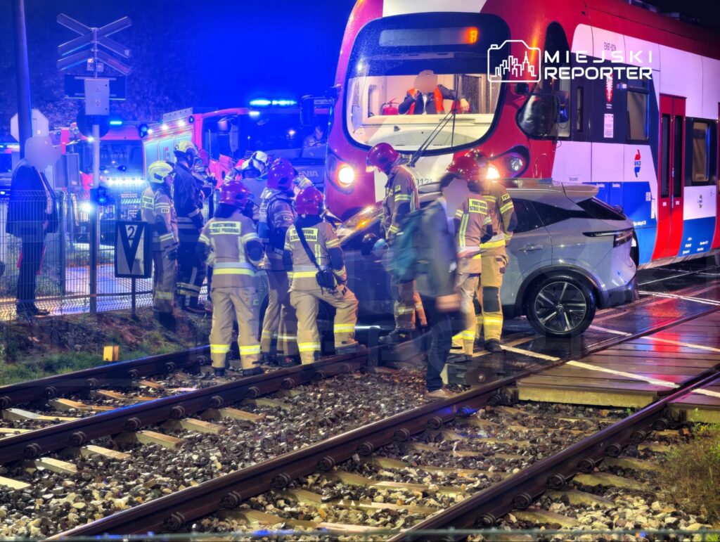 Strażacy i ratownicy przy samochodzie osobowym na torowisku przed czerwonym tramwajem, nocą przy niebieskich światłach służb.