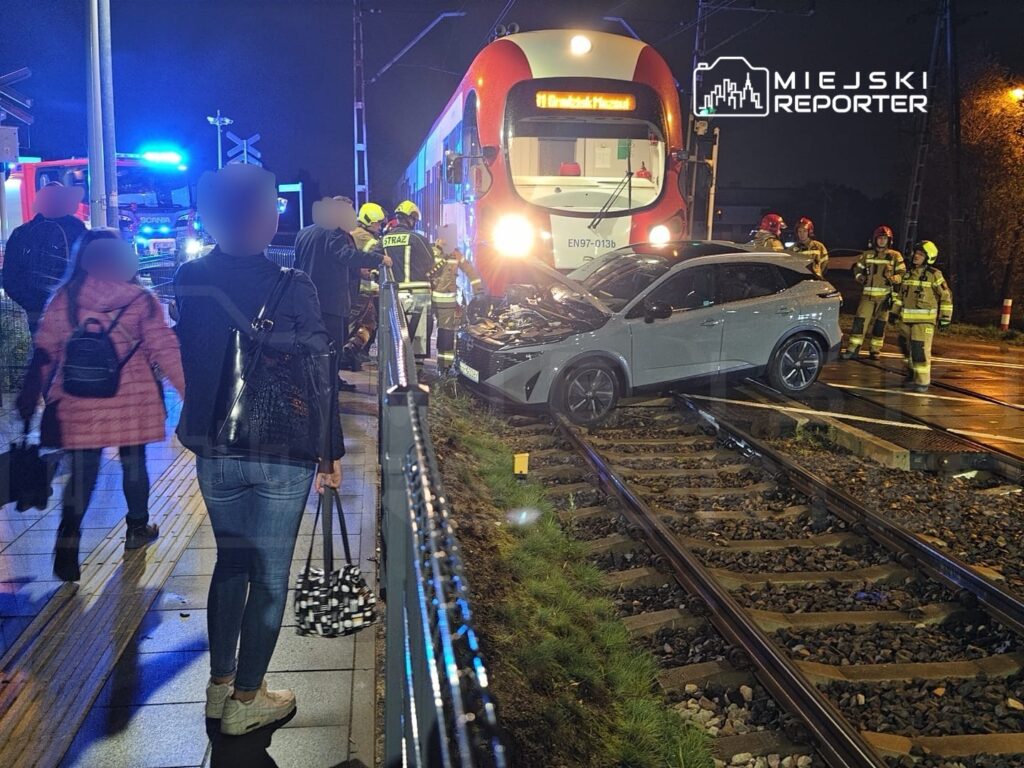 Samochód osobowy uszkodzony po zderzeniu z tramwajem na torowisku, obok strażacy i pasażerowie na peronie.