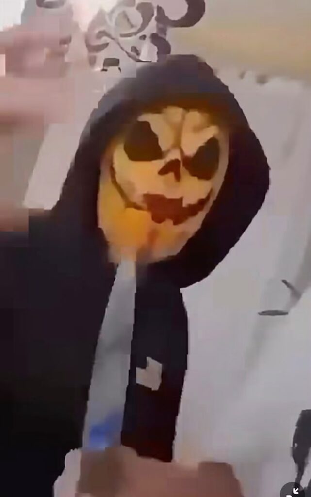 Osoba w czarnej bluzie z kapturem i maską przypominającą dynię stoi w pomieszczeniu.