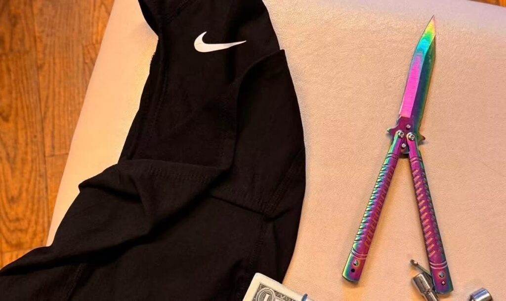 Czarne spodnie sportowe z logo Nike i kolorowy nóż motylkowy leżą na jasnej powierzchni obok drewnianej podłogi.