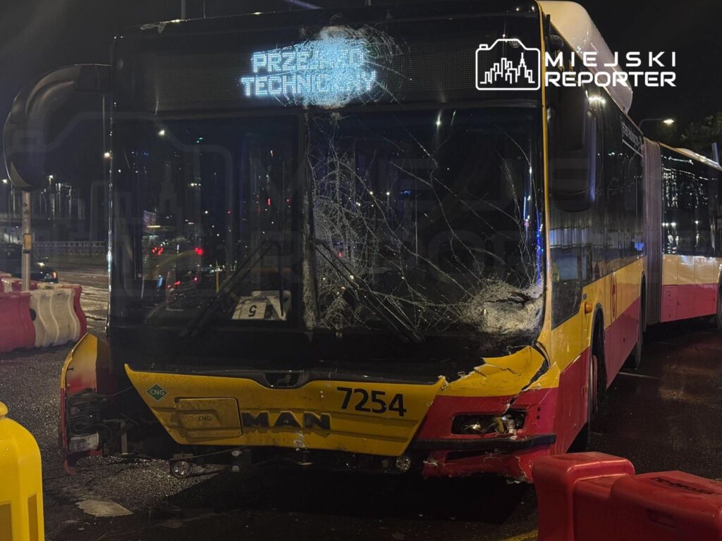 Uszkodzony żółto-czerwony autobus z rozbitą przednią szybą stoi nocą na ulicy, otoczony barierami drogowymi.