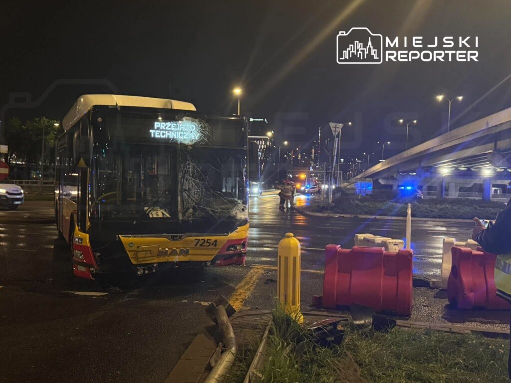 Uszkodzony żółty autobus miejski stoi nocą na mokrej jezdni przy barierach drogowych i wiadukcie.