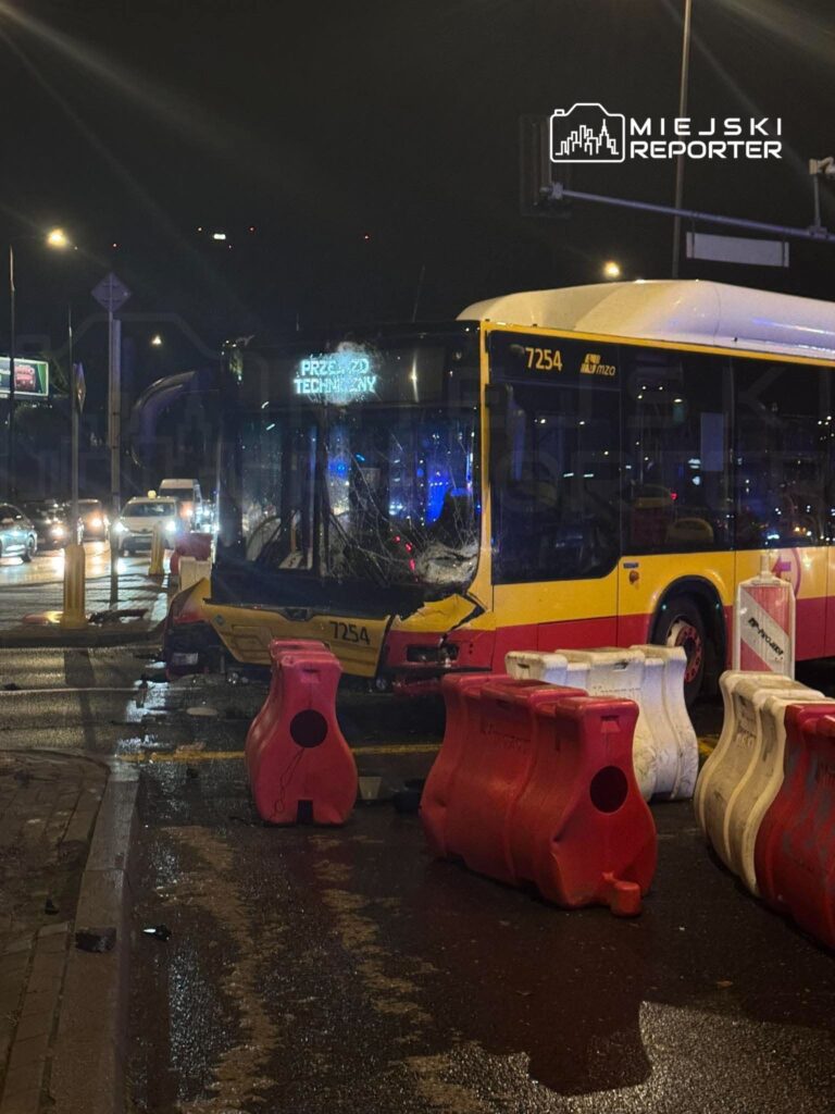 Uszkodzony żółto-czerwony autobus miejski z rozbitą przednią szybą stoi nocą na mokrej ulicy, za plastikowymi barierami.