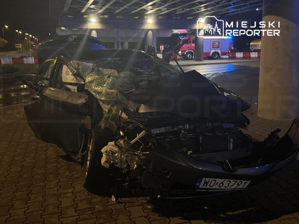 Rozbity samochód z uszkodzonym przodem i rozbitą szybą stoi nocą pod wiaduktem, w tle widać wóz strażacki.