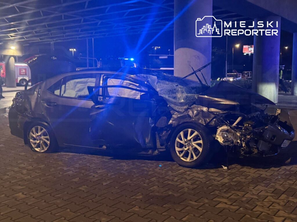 Rozbity samochód osobowy z mocno uszkodzonym przodem stoi nocą pod wiaduktem, oświetlony niebieskimi światłami.