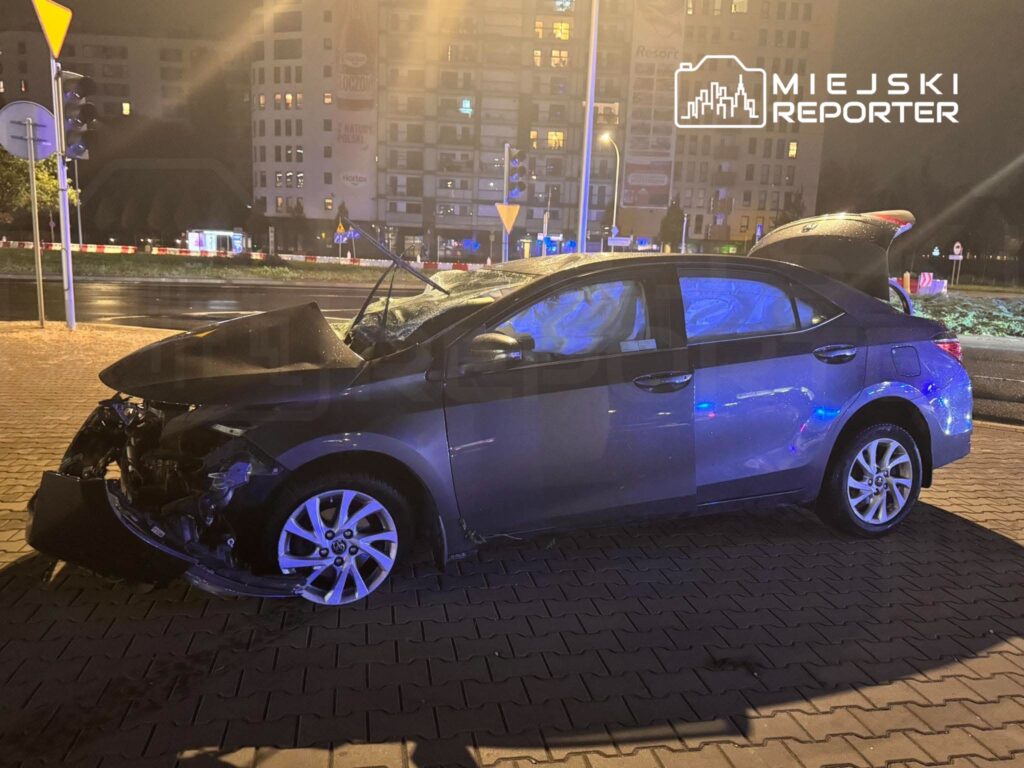 Uszkodzony szary sedan z podniesioną maską stoi nocą na chodniku przy miejskim skrzyżowaniu.