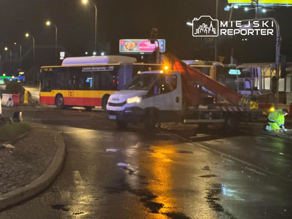 Ciężarówka z wysięgnikiem i autobus blokują nocną ulicę; pracownik w odblaskowym stroju klęczy przy jezdni.