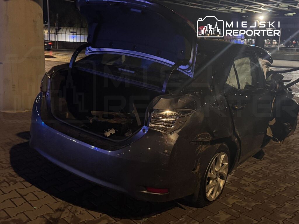 Uszkodzony szary samochód z otwartą klapą bagażnika stoi nocą na brukowanym parkingu.