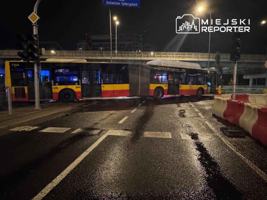 Przegubowy autobus miejski blokuje mokre skrzyżowanie nocą, pod wiaduktem i przy znakach drogowych.