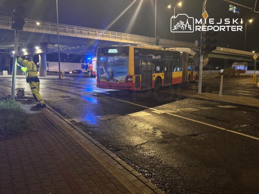 Autobus miejski stoi na mokrym skrzyżowaniu pod wiaduktem, obok pracownik w odblaskowym stroju i pojazd służb.