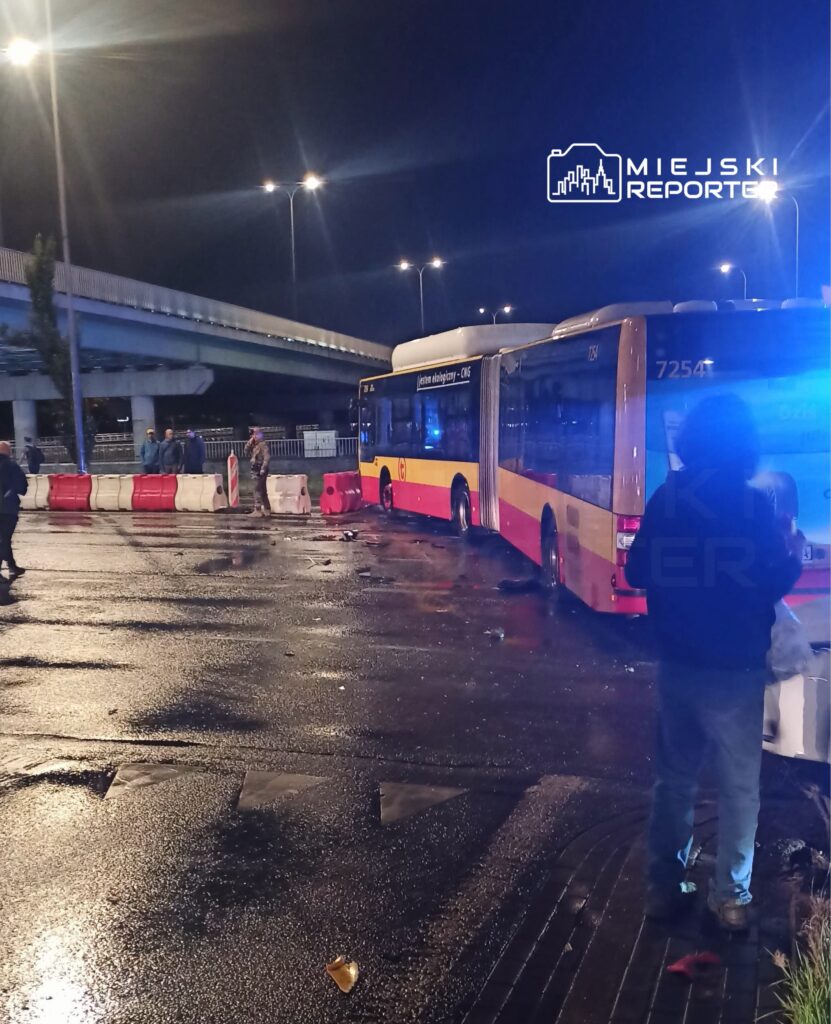 Przegubowy autobus miejski stoi nocą na mokrej jezdni przy barierach drogowych, oświetlony niebieskim światłem.