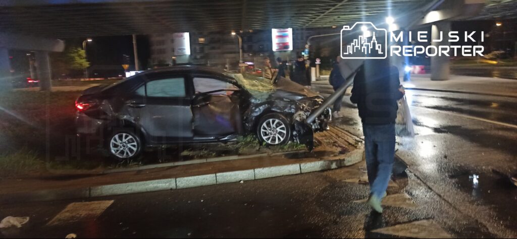 Uszkodzony samochód osobowy po kolizji stoi na wysepce pod wiaduktem, obok przechodzi mężczyzna w kamizelce odblaskowej.