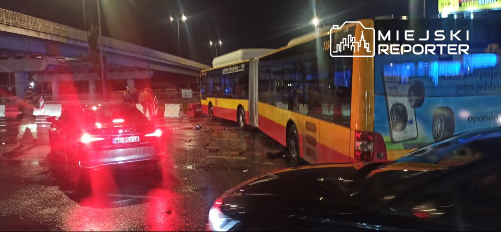 Autobus miejski i samochody stoją na mokrej jezdni nocą pod wiaduktem, oświetlone światłami pojazdów.