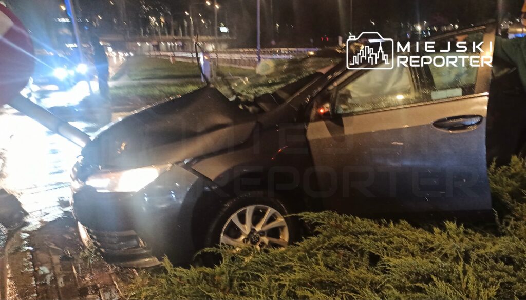 Uszkodzony samochód osobowy z otwartymi drzwiami stoi nocą na trawniku przy drodze, obok świecą światła pojazdu ratunkowego.