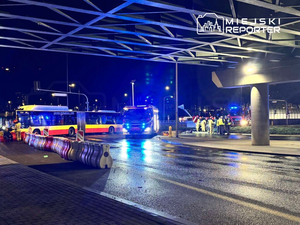 Autobusy stoją nocą na mokrej ulicy, a obok nich grupa służb w odblaskowych strojach pod zadaszeniem.