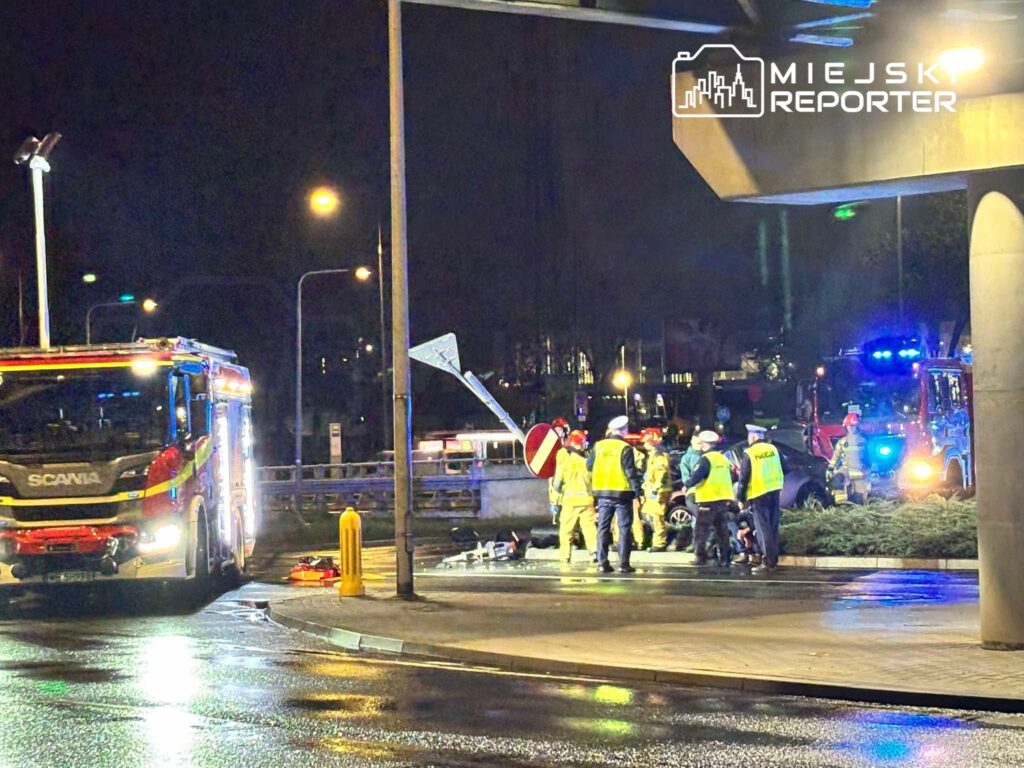 Służby ratunkowe w odblaskowych kamizelkach pracują nocą przy wypadku drogowym, obok stoją wozy strażackie i autobus.