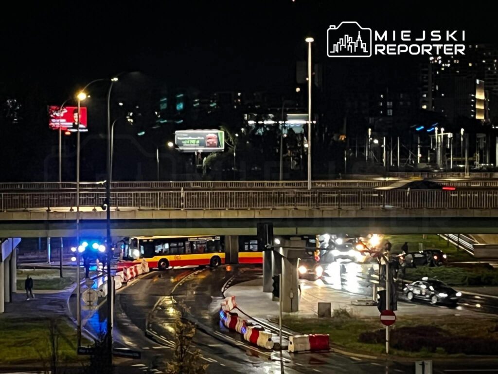 Autobus miejski i samochody jadą nocą pod wiaduktem, przy jezdni stoją bariery drogowe.
