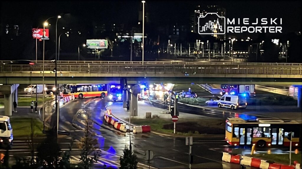 Służby ratunkowe i autobusy na nocnym skrzyżowaniu pod wiaduktem, przy zamkniętym odcinku drogi.