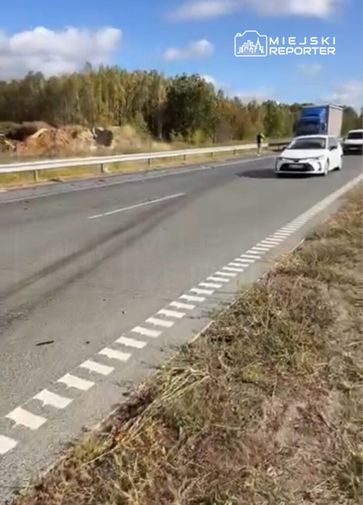 Biały samochód i ciężarówka jadą drogą z barierą, przy której stoi pieszy; w tle widać drzewa.