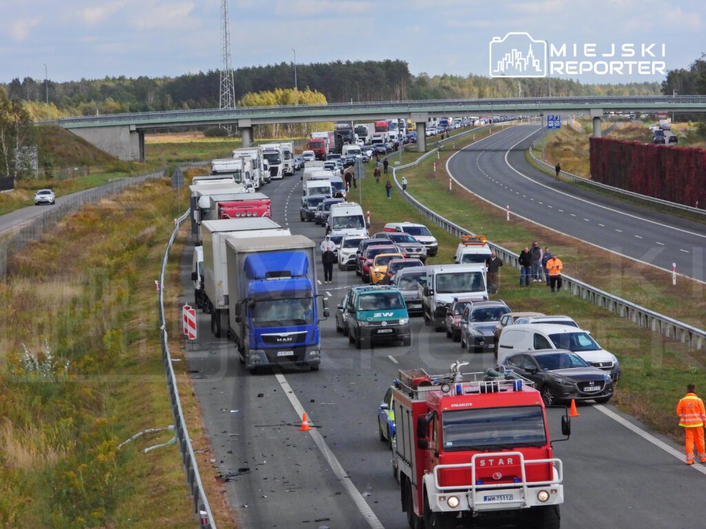 Korek ciężarówek i samochodów osobowych na autostradzie, obok pojazdy straży pożarnej i służby drogowe.