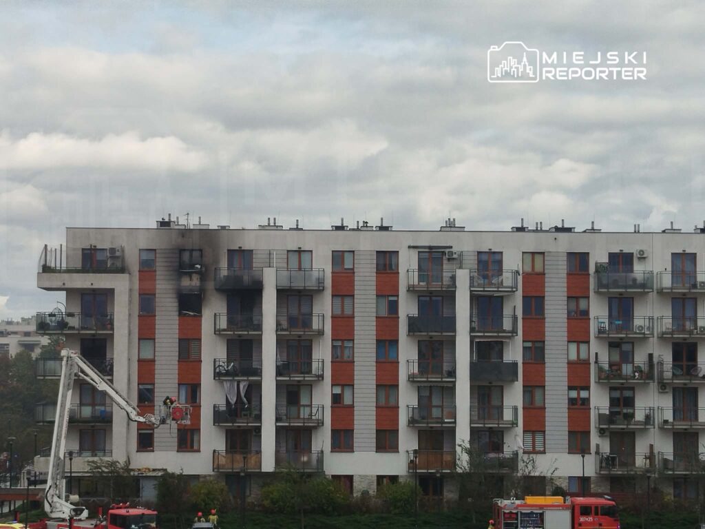 Wielopiętrowy blok mieszkalny z balkonami, przy którym stoją wozy strażackie i podnośnik koszowy.