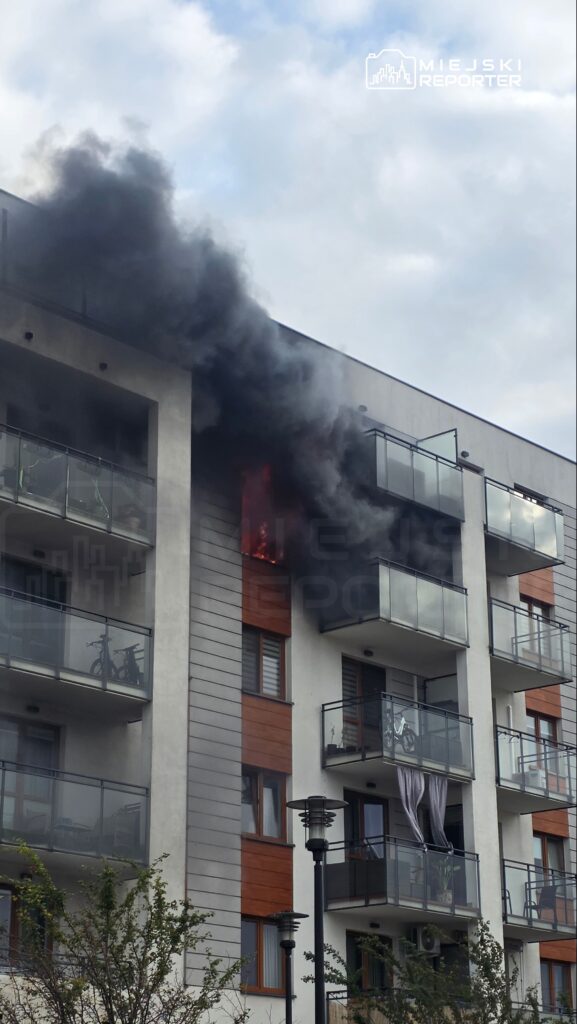 Płomienie i gęsty czarny dym wydobywają się z okna na piętrze bloku mieszkalnego.