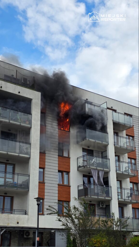 Ogień i gęsty, czarny dym wydobywają się z okna na piętrze bloku mieszkalnego z balkonami.