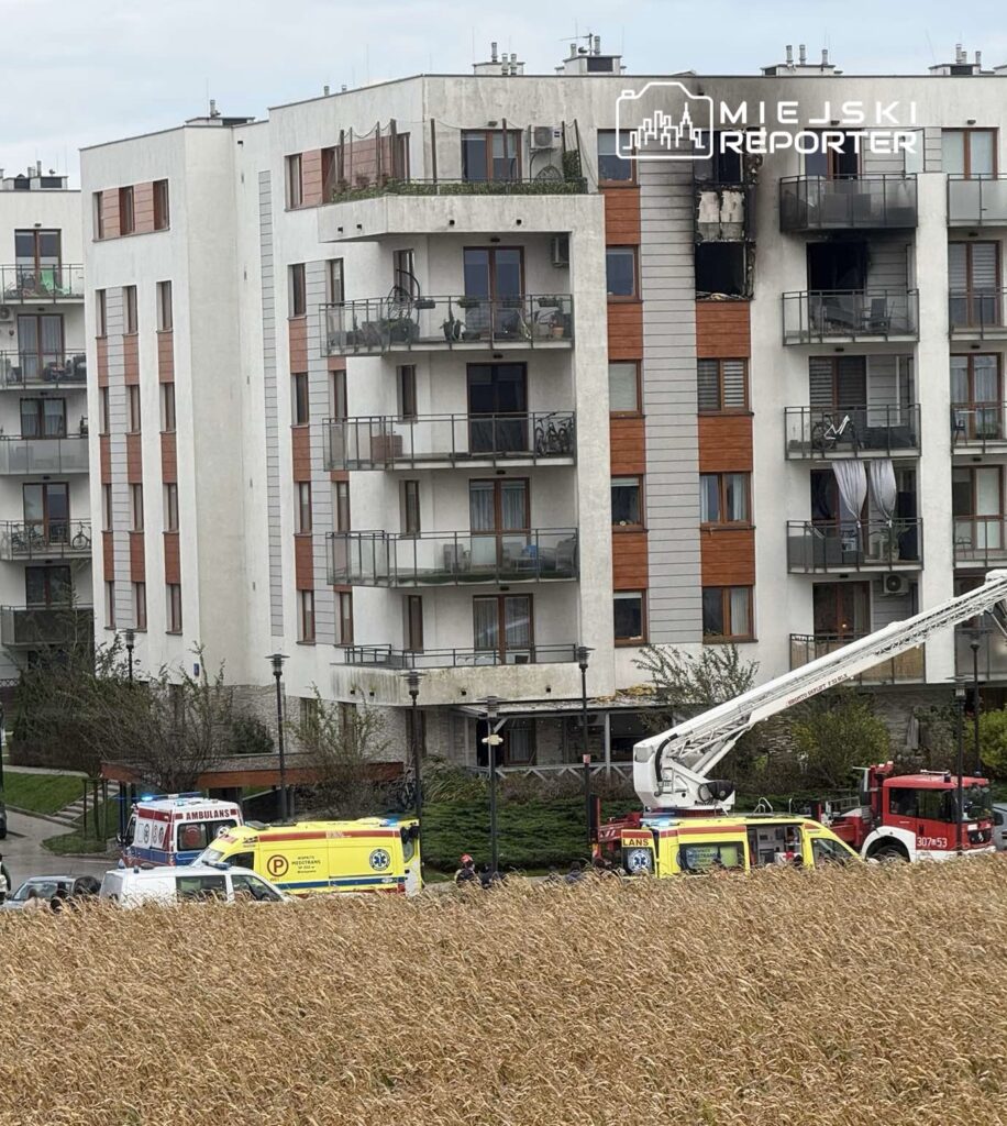 Strażacy i ratownicy medyczni interweniują przy bloku mieszkalnym; na miejscu stoją karetki i wóz strażacki z podnośnikiem.