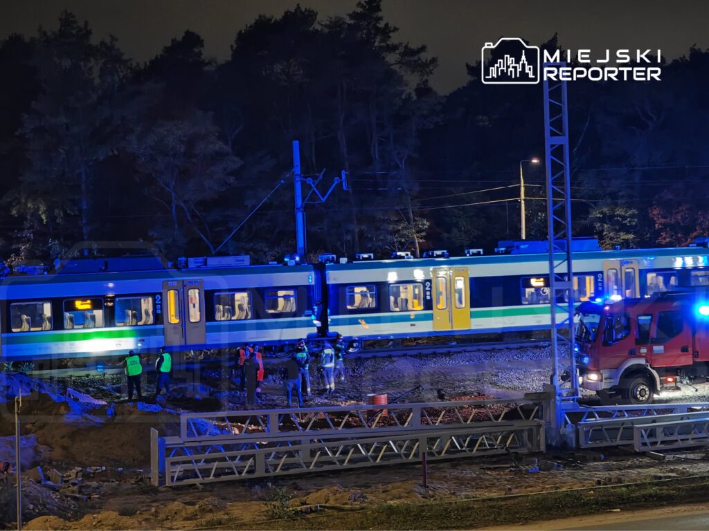 Pociąg pasażerski stoi nocą na torach, obok niego pracują służby ratunkowe i stoi wóz strażacki.