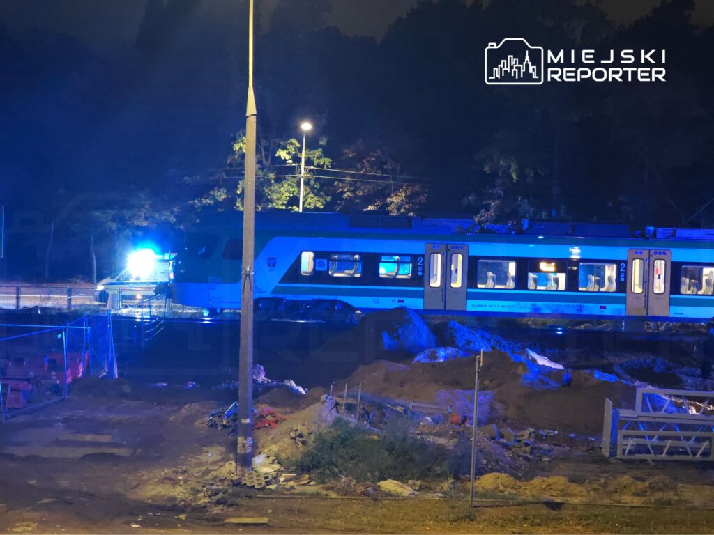 Pociąg pasażerski stoi nocą przy oświetlonym niebieskimi światłami miejscu robót ziemnych.