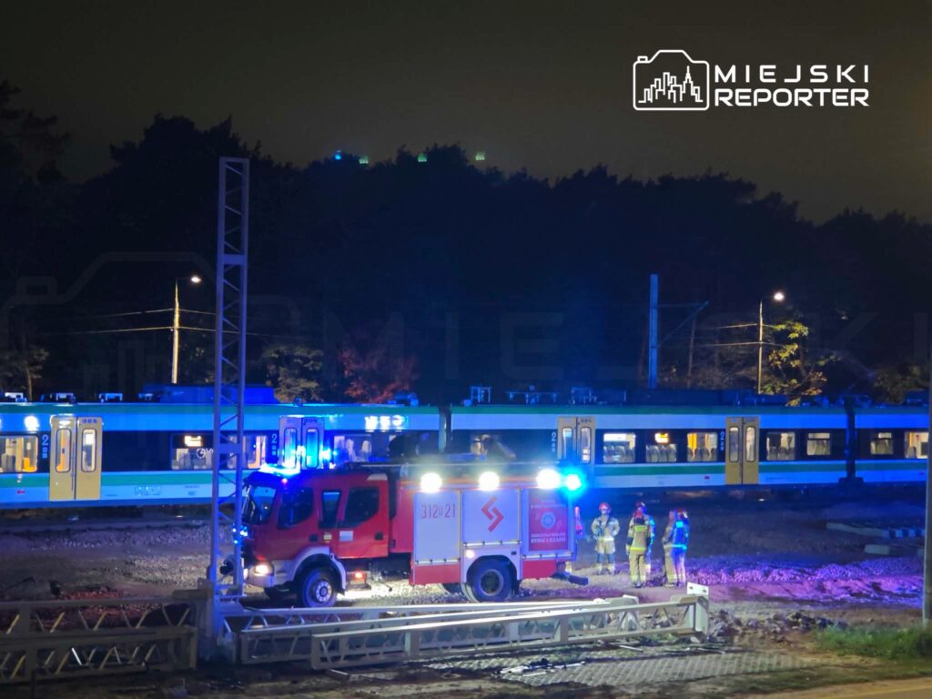 Strażacy stoją przy wozie strażackim obok niebiesko-białego pociągu na torach nocą.