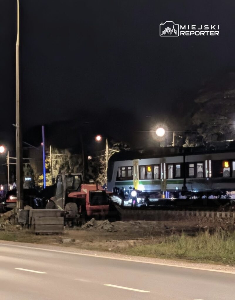 Pociąg stoi nocą przy torach obok czerwonej maszyny budowlanej i rozkopanego terenu.