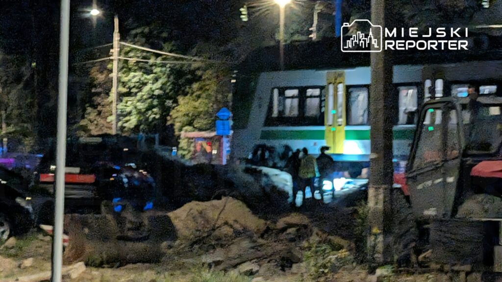 Nocą tramwaj i samochody stoją przy wykopie, a kilka osób obserwuje miejsce zdarzenia.