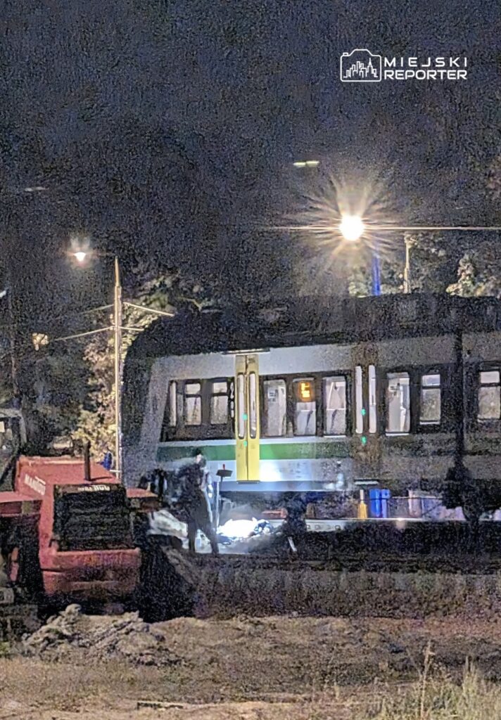 Tramwaj stoi nocą na torowisku obok czerwonego pojazdu technicznego, przy oświetlonej ulicy.