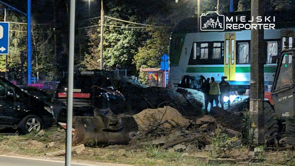 Uszkodzony samochód leży na boku przy torowisku, obok stoją tramwaj i kilka osób nocą.