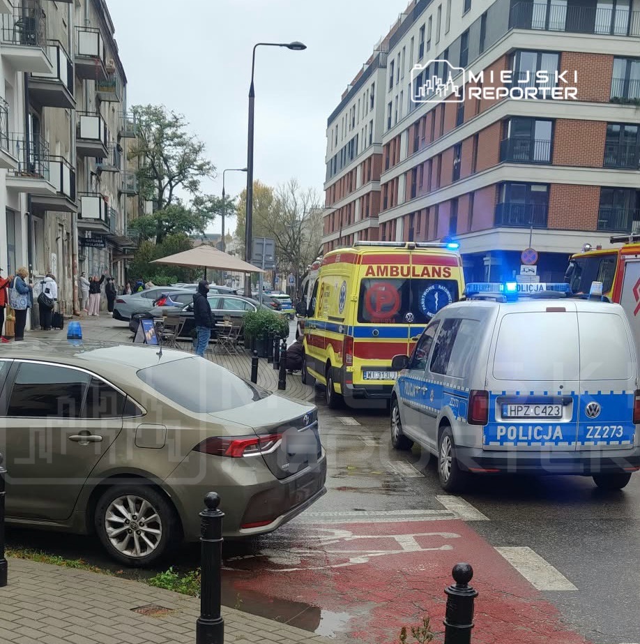 Radiowóz policji i karetka z włączonymi sygnałami stoją na ulicy między budynkami mieszkalnymi.