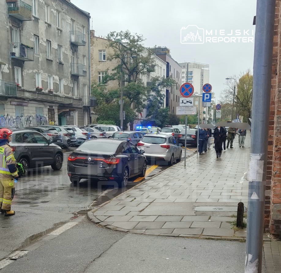 Samochody policyjne z włączonymi niebieskimi światłami stoją przy ulicy, obok nich widać funkcjonariuszy i strażaka.