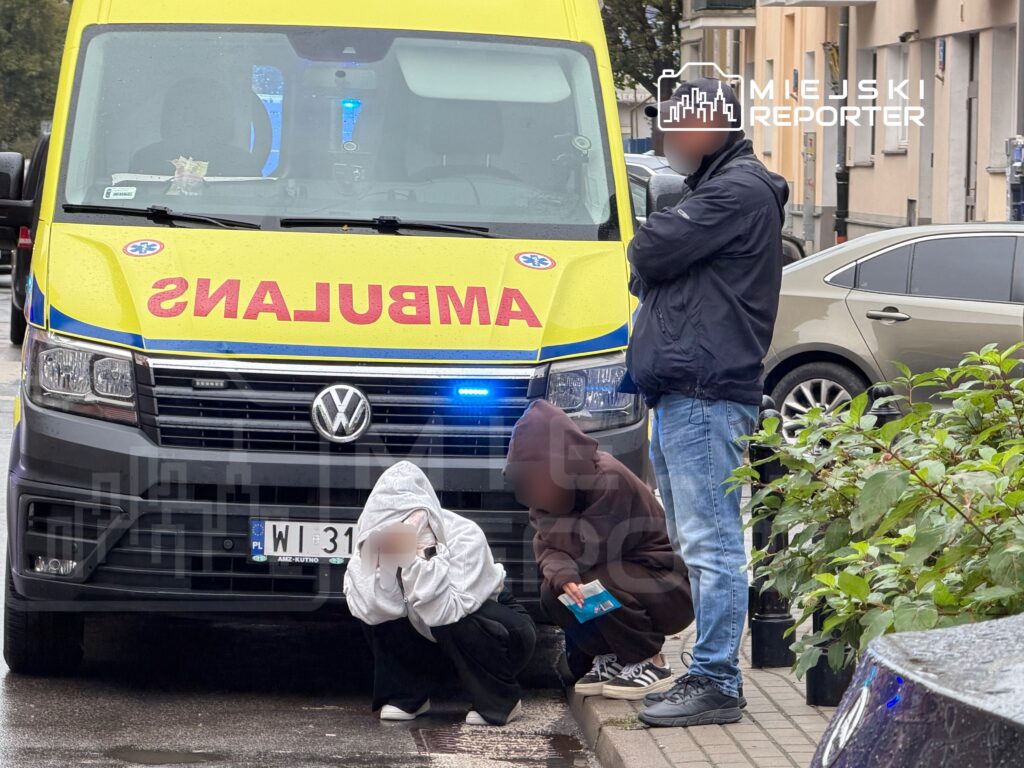 Ratownicy medyczni stoją przy karetce, a dwie osoby siedzą na chodniku przed budynkiem mieszkalnym.