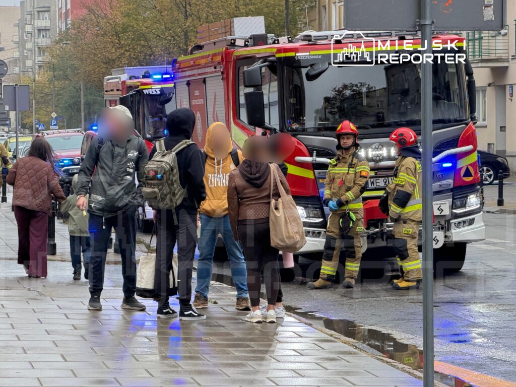 Strażacy stoją przy wozie strażackim, a kilka osób rozmawia z nimi na mokrym chodniku.