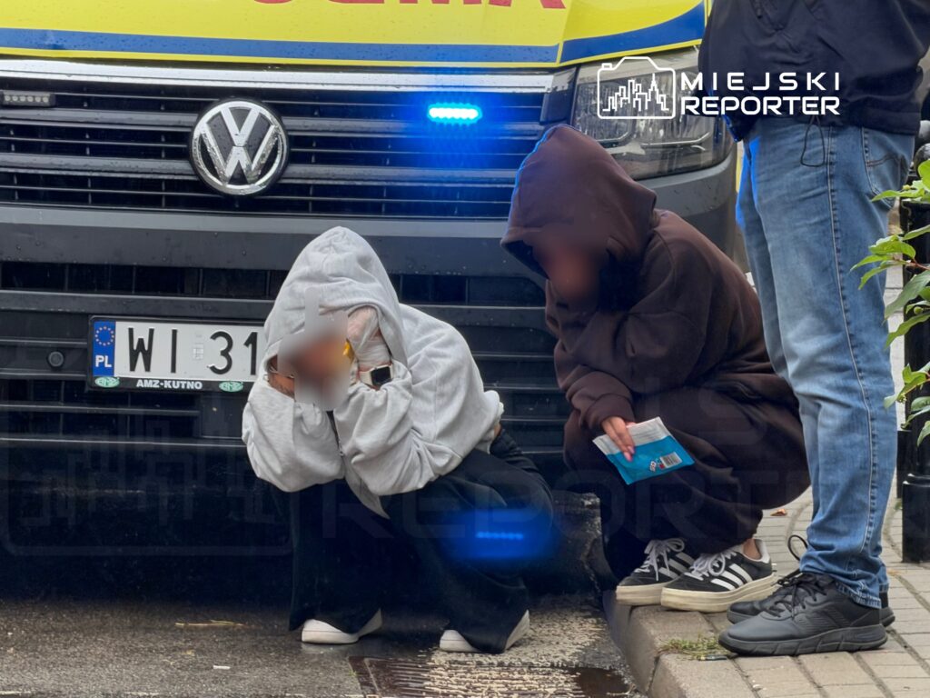Dwóch zakapturzonych młodych ludzi kuca przed pojazdem z niebieskimi światłami, obok stojącego mężczyzny w dżinsach.