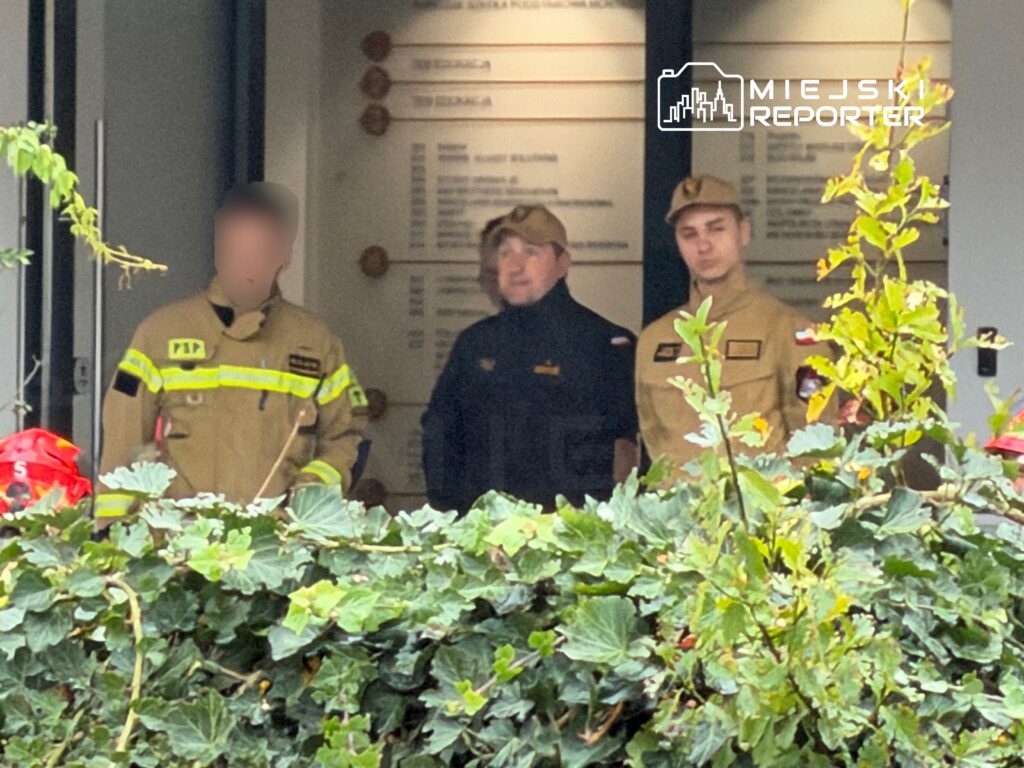Dwóch strażaków w mundurach i mężczyzna w czarnej kurtce stoją w wejściu do budynku za krzewami.