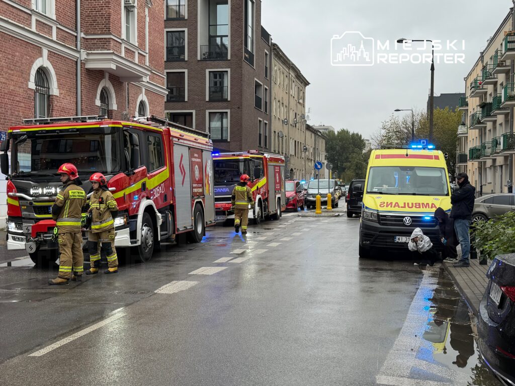 Strażacy stoją przy wozach strażackich, a obok na mokrej ulicy parkuje karetka pogotowia z włączonymi światłami.