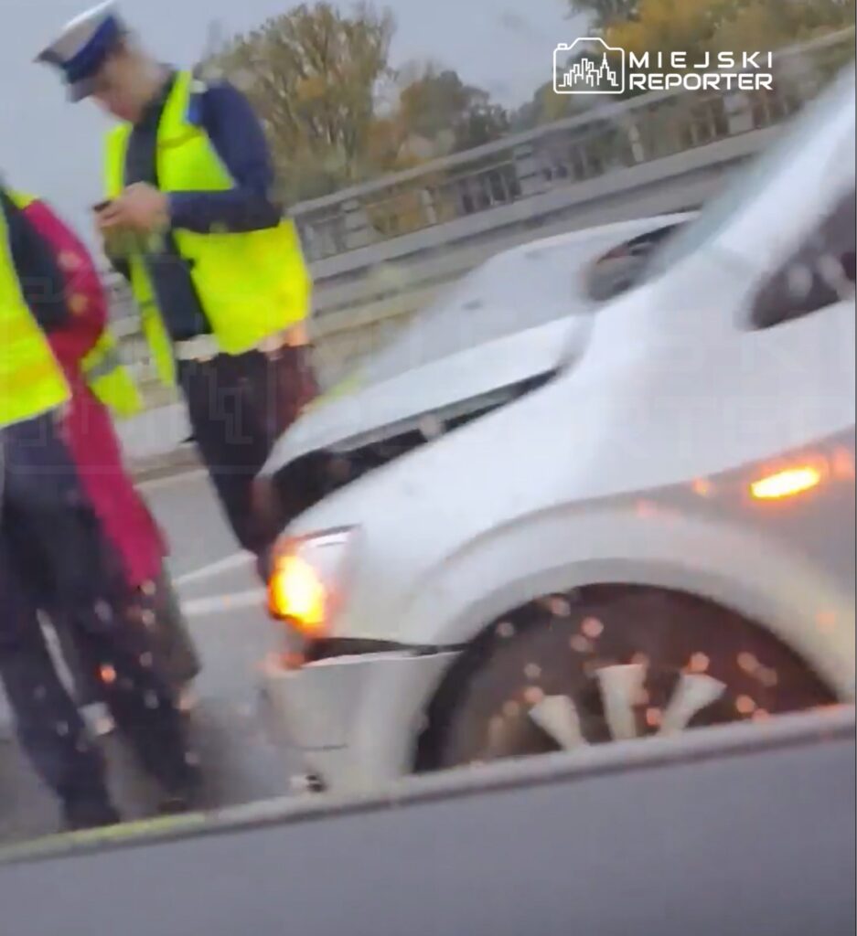 Policjanci w odblaskowych kamizelkach stoją obok białego samochodu na jezdni przy barierze mostu.