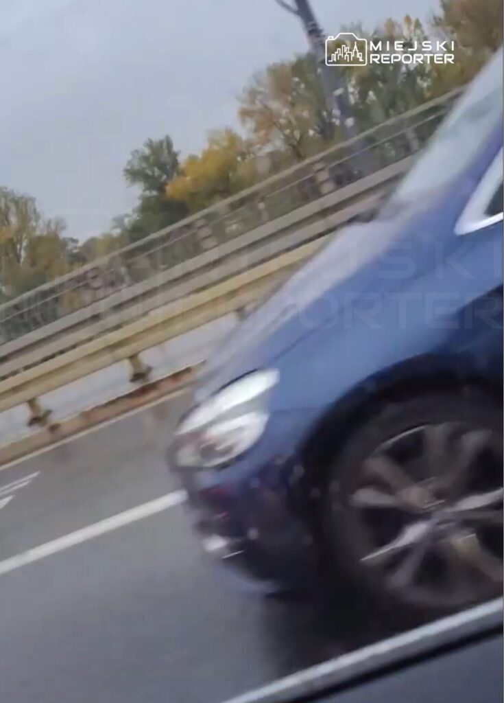 Niebieski samochód osobowy jedzie po jezdni obok barier mostu, w tle widać drzewa.