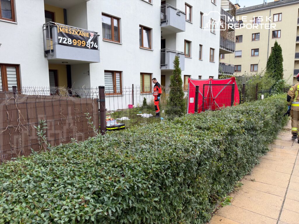 Ratownik w czerwonym stroju stoi przy bloku mieszkalnym obok czerwonego namiotu i ogrodzonego ogródka.