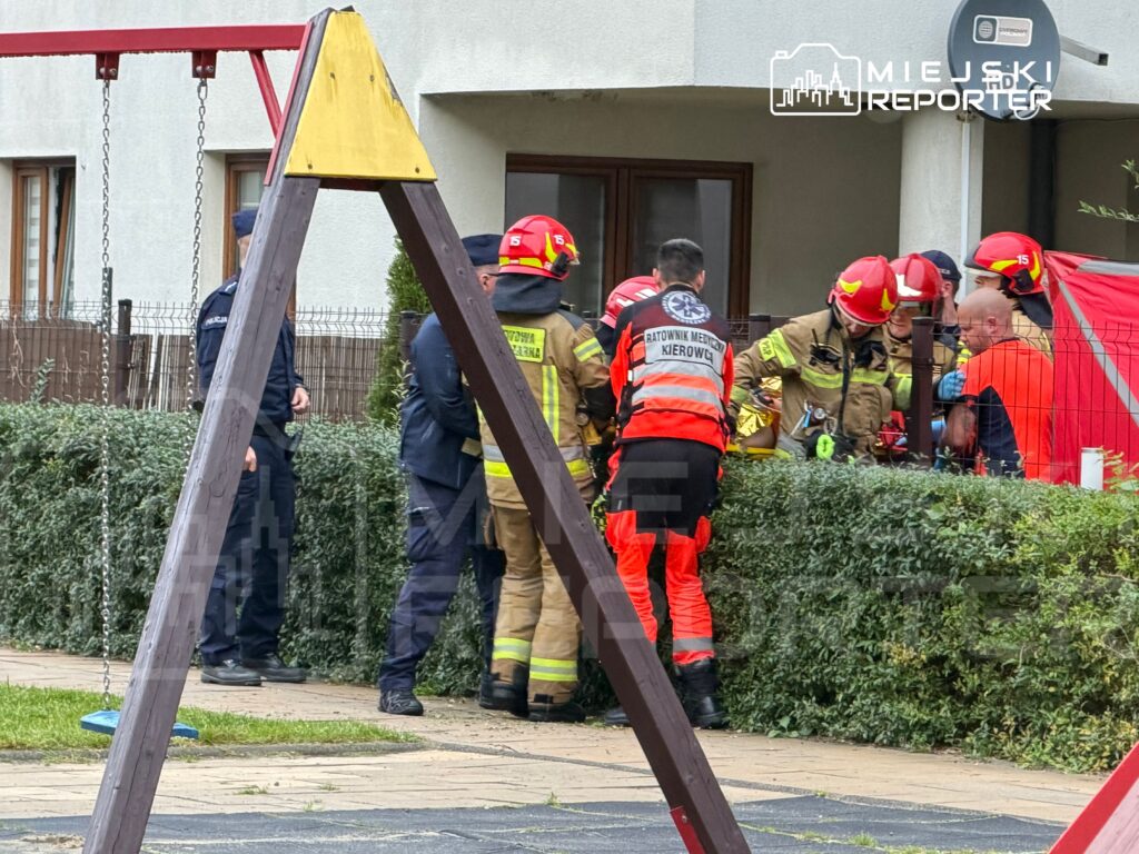 Strażacy i ratownicy medyczni zebrani przy wejściu do budynku mieszkalnego, za żywopłotem i ogrodzeniem.