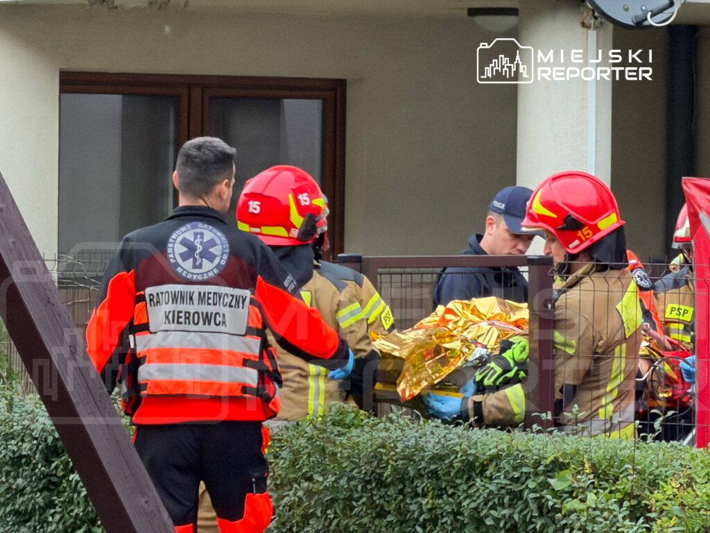 Ratownicy medyczni i strażacy przenoszą osobę okrytą złotą folią przed wejściem do budynku.