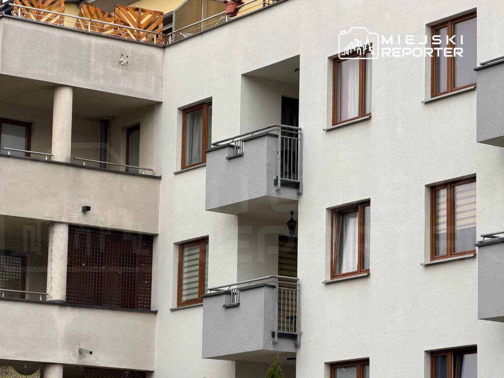 Jasna elewacja wielorodzinnego budynku mieszkalnego z balkonami, metalowymi balustradami i oknami w drewnianych ramach.