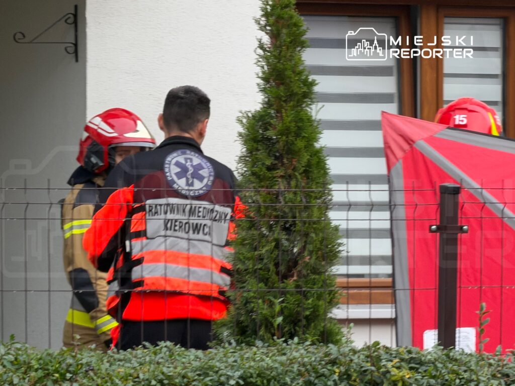Ratownik medyczny i strażacy w kaskach stoją za ogrodzeniem przed budynkiem, obok czerwonego parawanu.